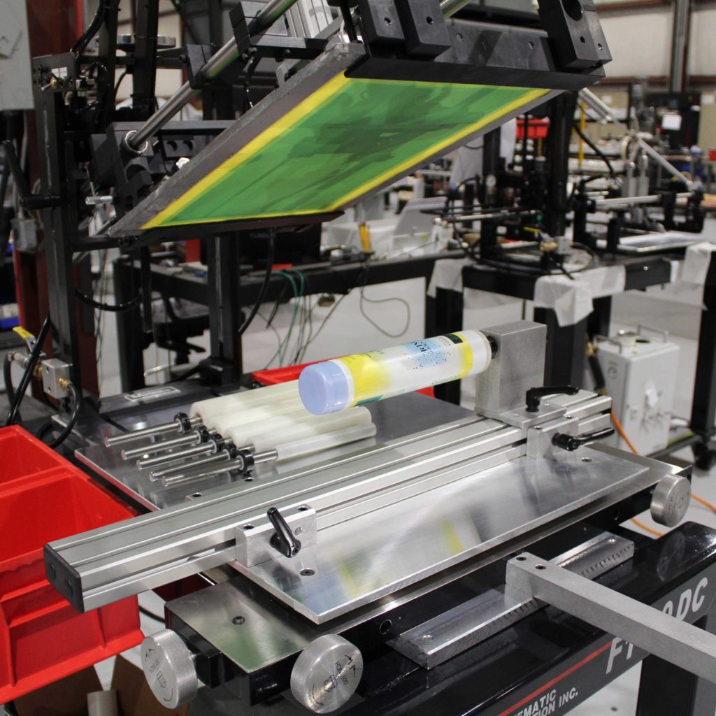 A screen printing machine in a factory setting printing a colorful design onto a cylindrical plastic container