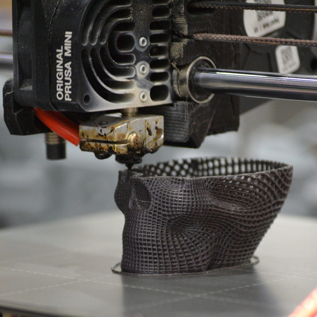 A close-up of a 3D printer nozzle fabricating a black mesh-like skull model using filament on a Prusa Mini printer