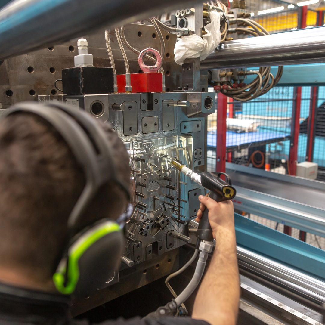 A worker wearing protective headphones uses a tool to clean or inspect a complex metal mold inside an industrial injection molding machine