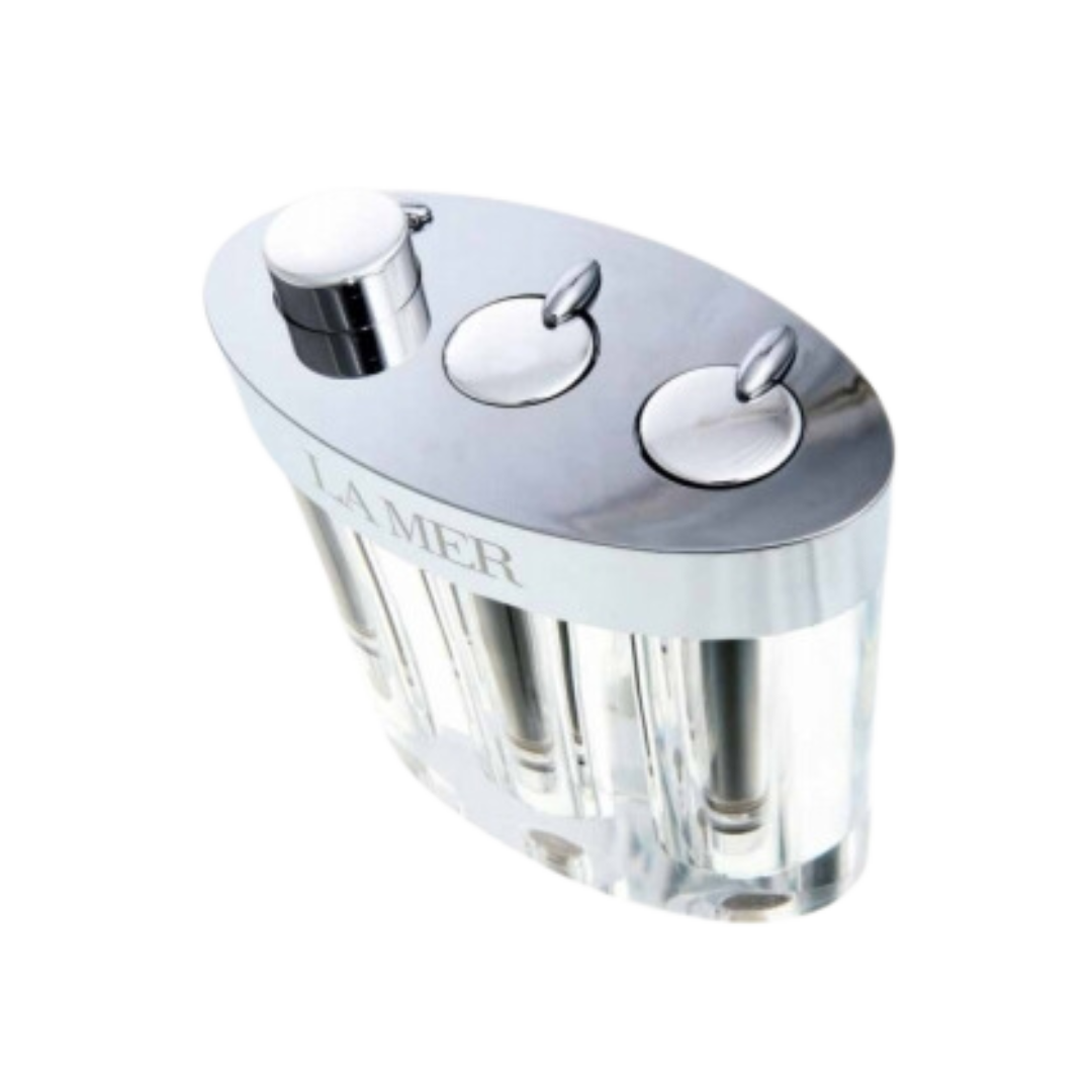 A sleek, silver-toned La Mer skincare set featuring three cylindrical containers housed in a clear, oval acrylic base with metallic caps and pump dispensers
