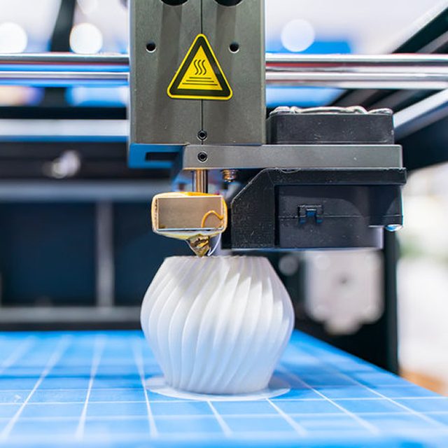 A 3D printer in the process of printing a white, spiral-patterned vase on a blue build platform
