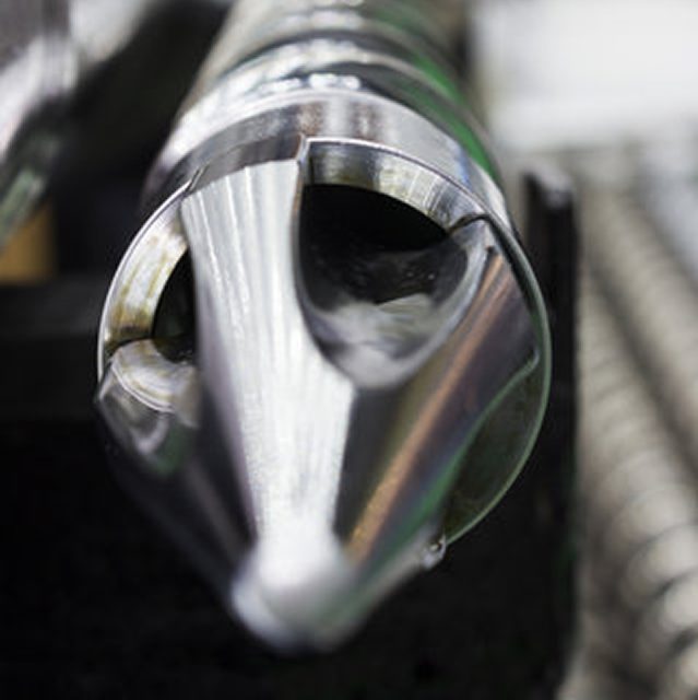 Close-up of a polished, metallic injection molding screw tip, used in plastic manufacturing machinery, with helical grooves visible in the blurred background