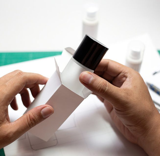 A person holding a white cosmetic bottle with a black cap, partially removed from a plain white box, with additional packaging materials in the background