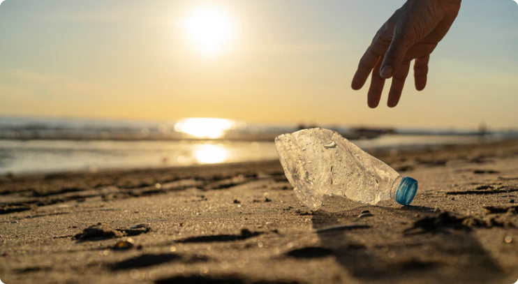 A hand reaches down to pick up a discarded plastic bottle lying on a sandy beach during sunset, with sunlight reflecting off the ocean in the background