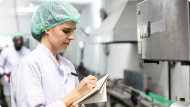 A woman in a lab coat and hairnet takes notes on a clipboard while inspecting equipment in a clean, industrial production facility