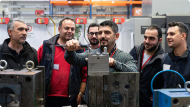 A group of male workers gathers around a mold tool in a workshop, with one man demonstrating or adjusting the equipment while others observe attentively