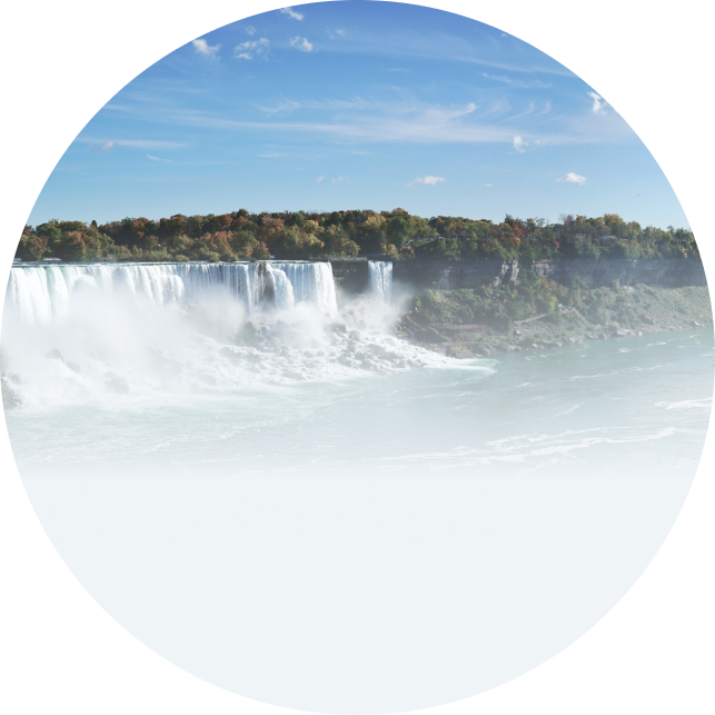 A scenic view of Niagara Falls with powerful waterfalls cascading over the edge, backed by a forested landscape under a bright blue sky