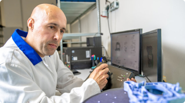 A technician in a lab coat operates a 3D scanning device while analyzing a blue mechanical part on a turntable, with CAD models displayed on dual monitors