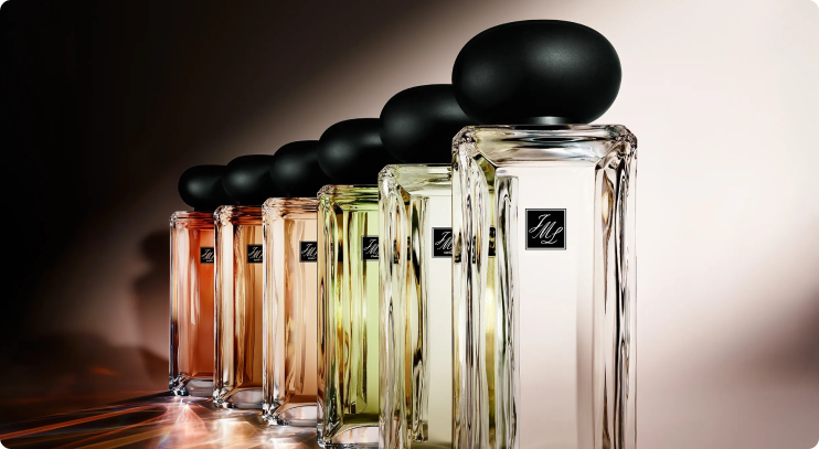 A row of elegant Jo Malone perfume bottles in various pastel shades with black rounded caps, arranged diagonally on a reflective surface
