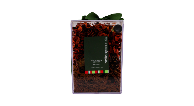 A clear rectangular gift box filled with dried botanicals labeled "Holiday Secrets" and "Sensuous Spruce Potpourri" by Victoria's Secret, topped with a dark green ribbon