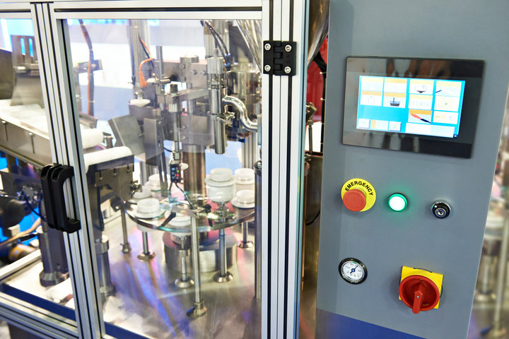 Automated packaging machine with a transparent enclosure, visible rotating platform holding white plastic containers, and a control panel featuring a touchscreen interface, buttons, and emergency stop controls