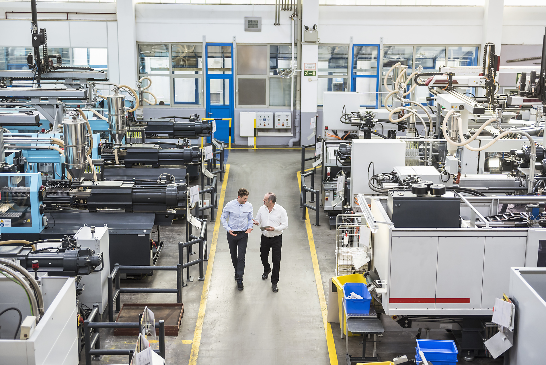 Two men walk and talk down the central aisle of a brightly lit, modern factory filled with large industrial machines and robotic arms