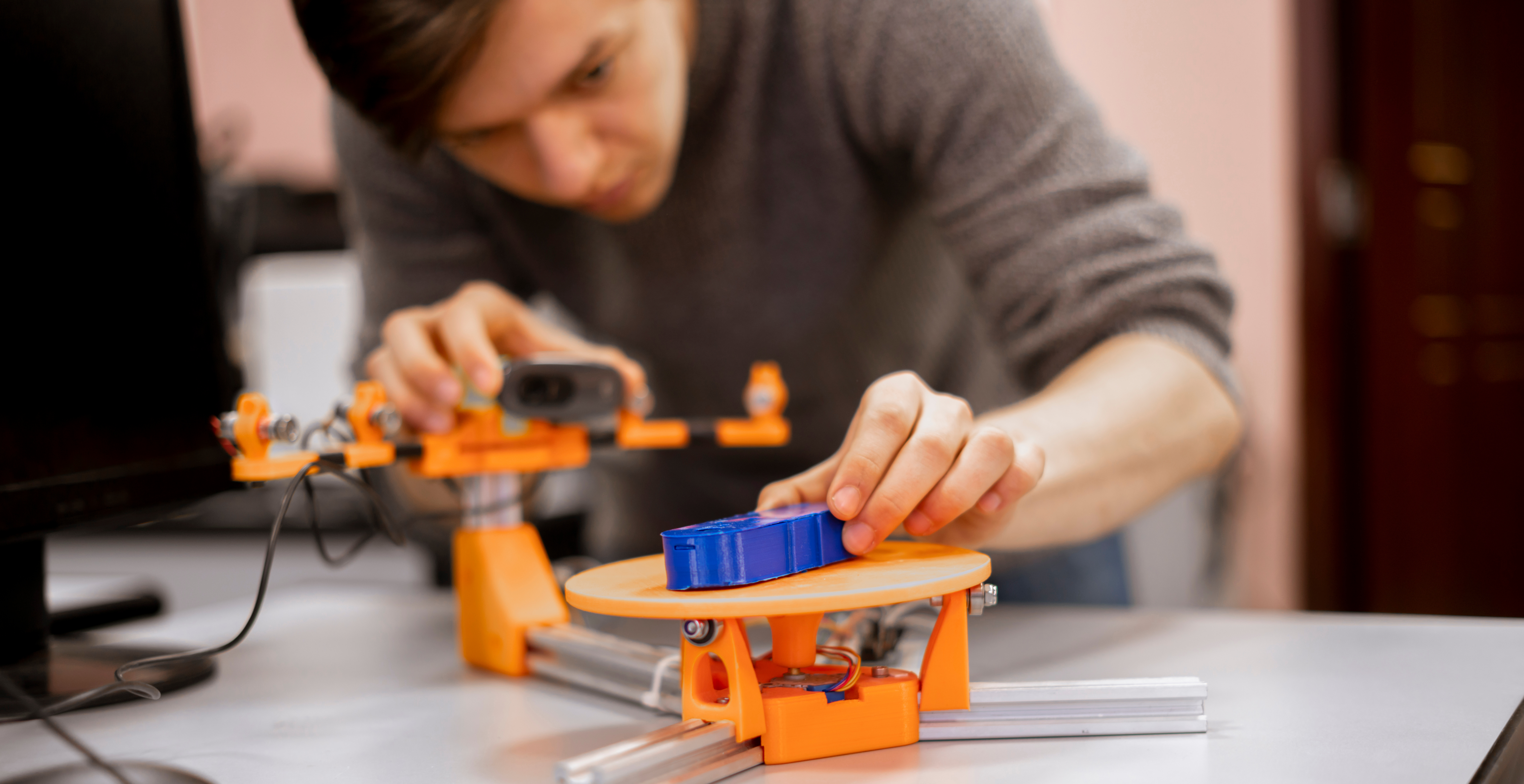 A person carefully places a blue 3D-printed object onto the scanning platform of an orange desktop 3D scanner setup