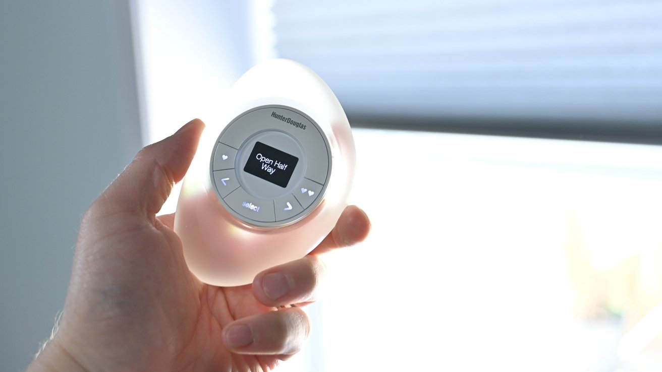 A person holding a Hunter Douglas remote control with a circular button interface and digital screen displaying the message “Open Half Way,” used for adjusting window blinds