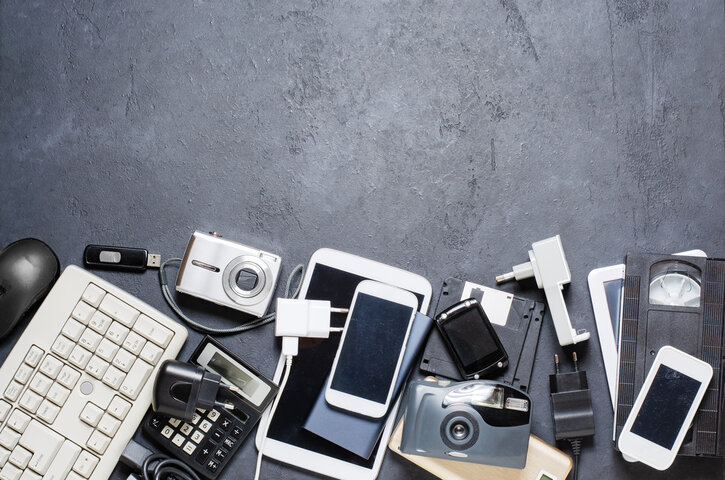 A collection of old electronic devices and accessories, including a keyboard, digital cameras, cell phones, chargers, a calculator, a USB stick, and VHS tapes, arranged on a dark gray surface