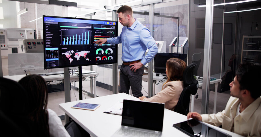 A man in a blue shirt presents analytics and data visualizations on a large screen to a group of colleagues seated around a conference table in a modern office setting