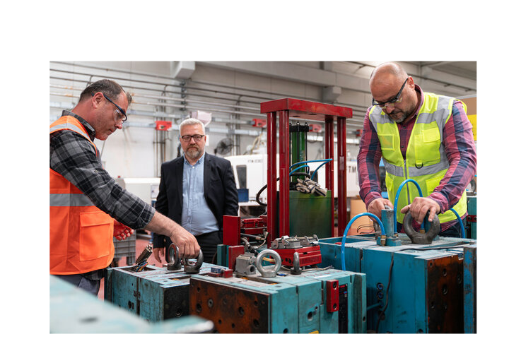 Two workers in safety vests operate heavy-duty mold components on a factory floor while a supervisor in business attire observes