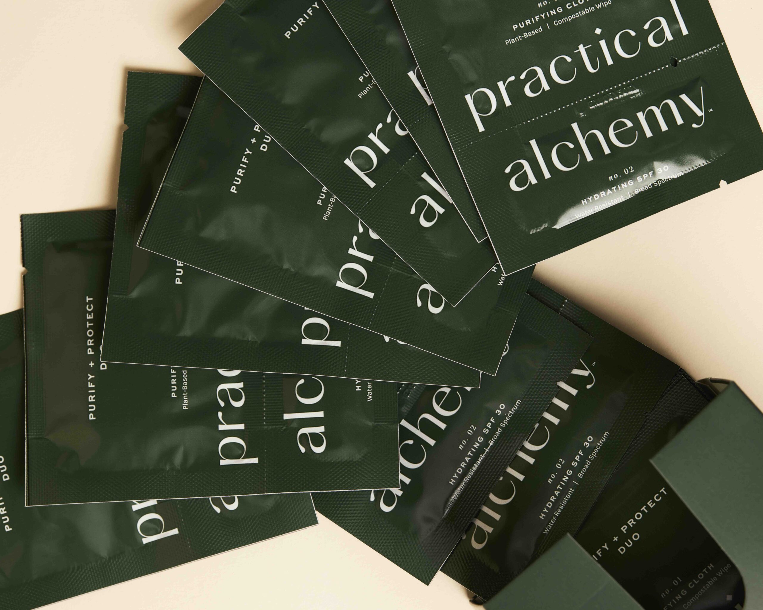 Multiple dark green sachets of "Practical Alchemy" purifying cloths are spread out on a beige surface, with some partially inside a matching green box. The packaging features minimalist white text highlighting plant-based ingredients and SPF30 hydration.